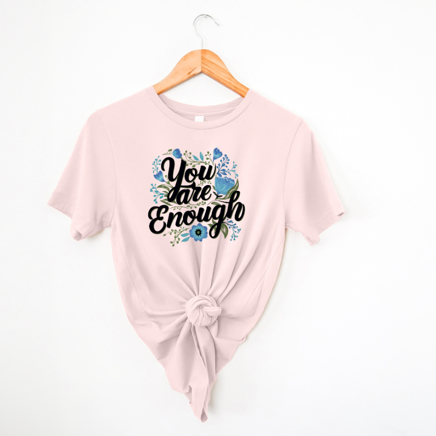 A pink colored t-shirt with the text 'You are Enough' printed on the front, surrounded by a floral design, displayed on a hanger.